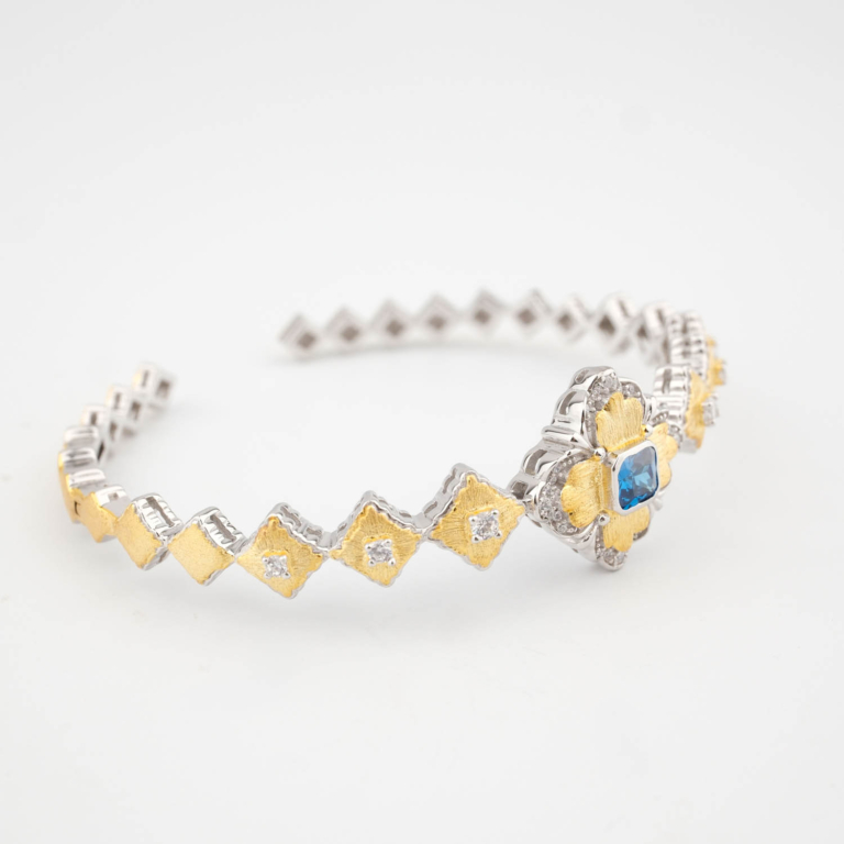 A luxury designer bangle bracelet featuring a square faceted blue gemstone surrounded by textured gold plated baroque details in 925 sterling silver.
