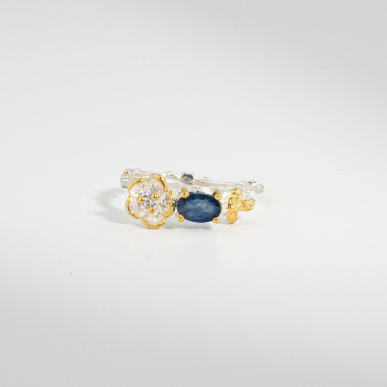 A 925 sterling silver women's ring featuring a natural oval blue sapphire gemstone accented with a gold-plated silver flower motif.